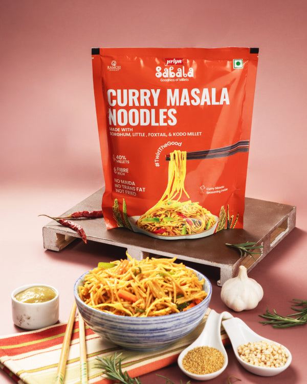 Millet Curry Masala Noodles pack with a bowl of spiced noodles, garlic, spices, and herbs on a wooden tray