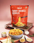 Millet Curry Masala Noodles pack with a bowl of spiced noodles, garlic, spices, and herbs on a wooden tray